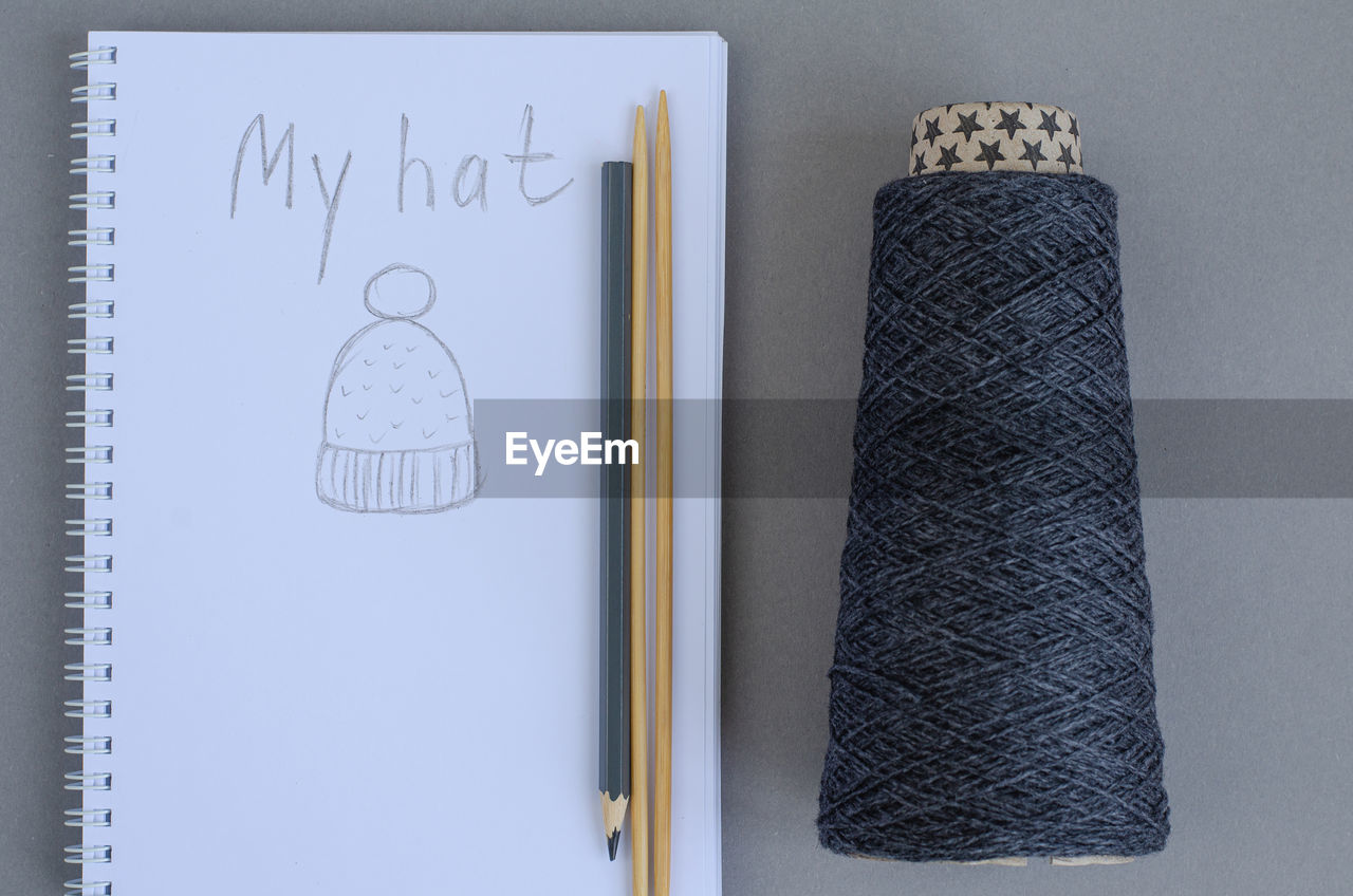 Dark gray yarn ball, bamboo knitting needles, pencil and notebook with sketch hat. gray background. 