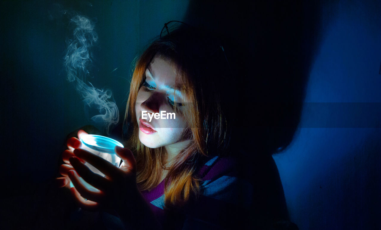 Close-up of woman with illuminated jar with smoke sitting in dark room