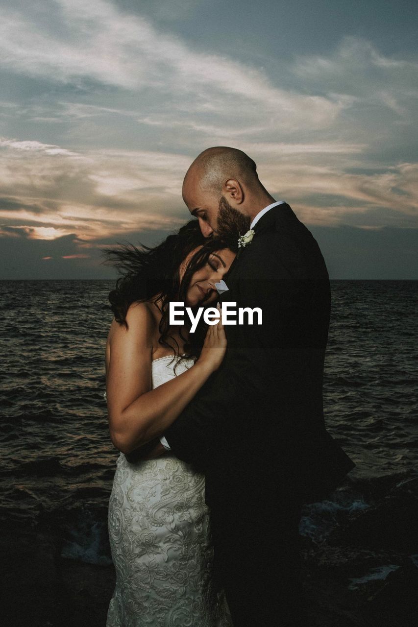 REAR VIEW OF MAN AND WOMAN STANDING AT SEA AGAINST SKY