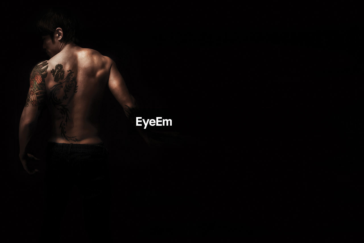 Rear view of young man with tattoo standing against black background