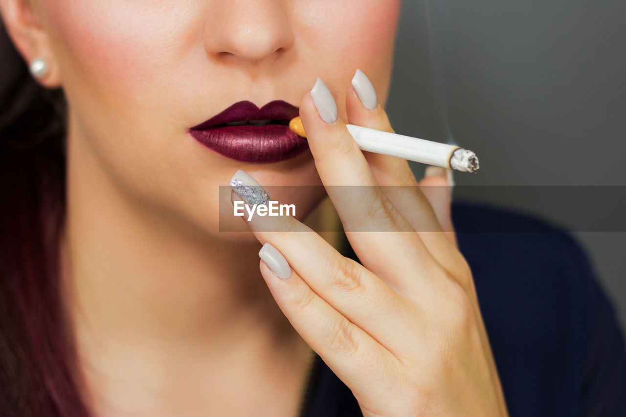 Midsection of young woman smoking cigarette against gray background