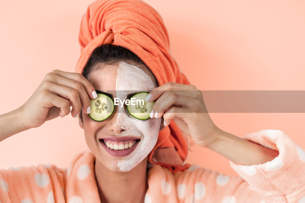 Close-up of smiling woman with facial mask holding cucumber slices on eyes against colored background