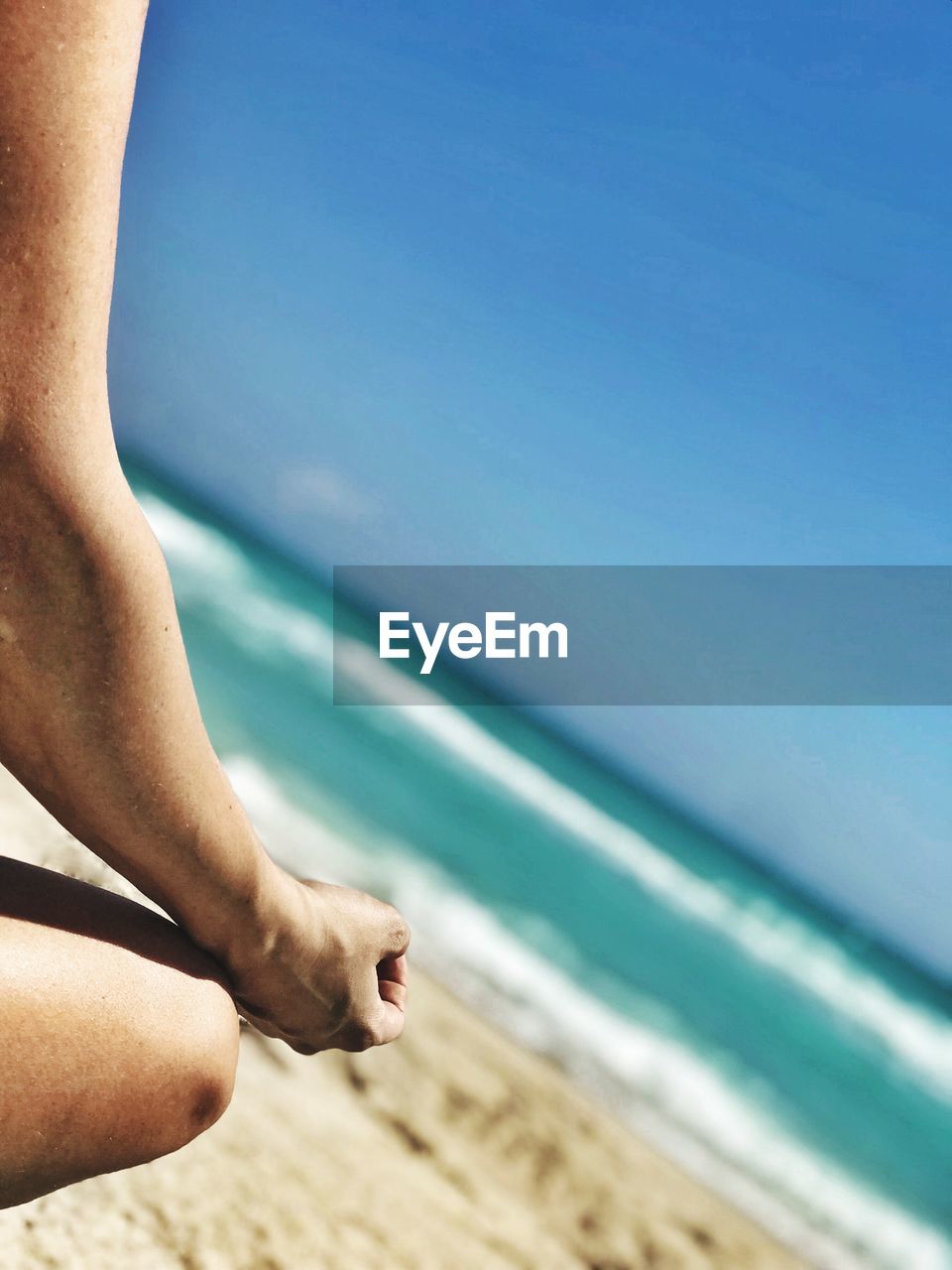 Midsection of woman meditating while sitting at beach against blue sky during sunny day