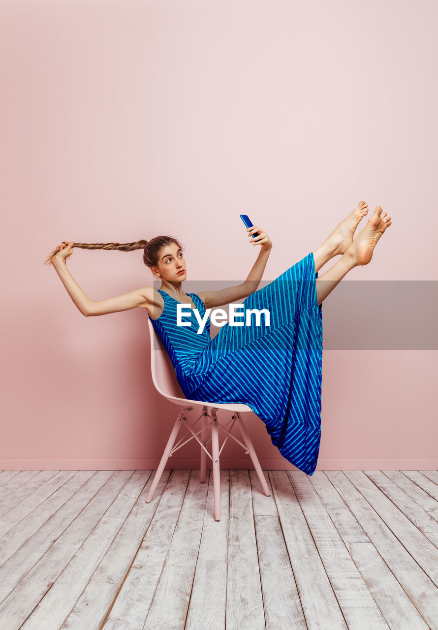 Side view of woman sitting in a chair lifting legs up barefoot looking at mobile phone while taking a selfie holding ponytail up on pink background