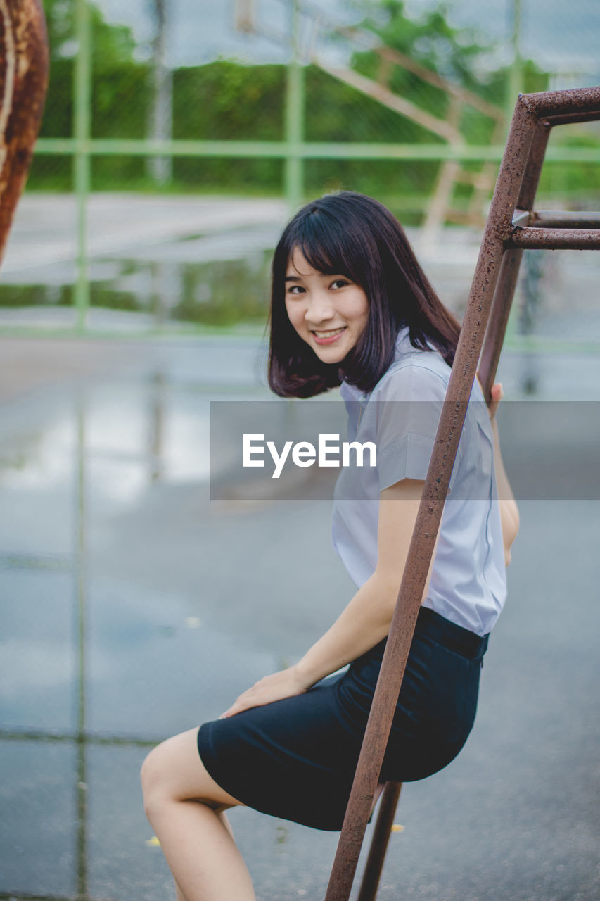 Portrait of smiling young woman sitting on metallic outdoor play equipment at playground during rainy season