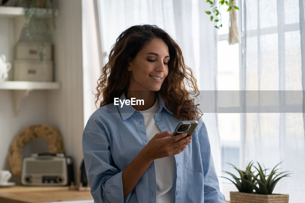 Positive smiling woman in casual clothes stands with phone in hand in house kitchen interior