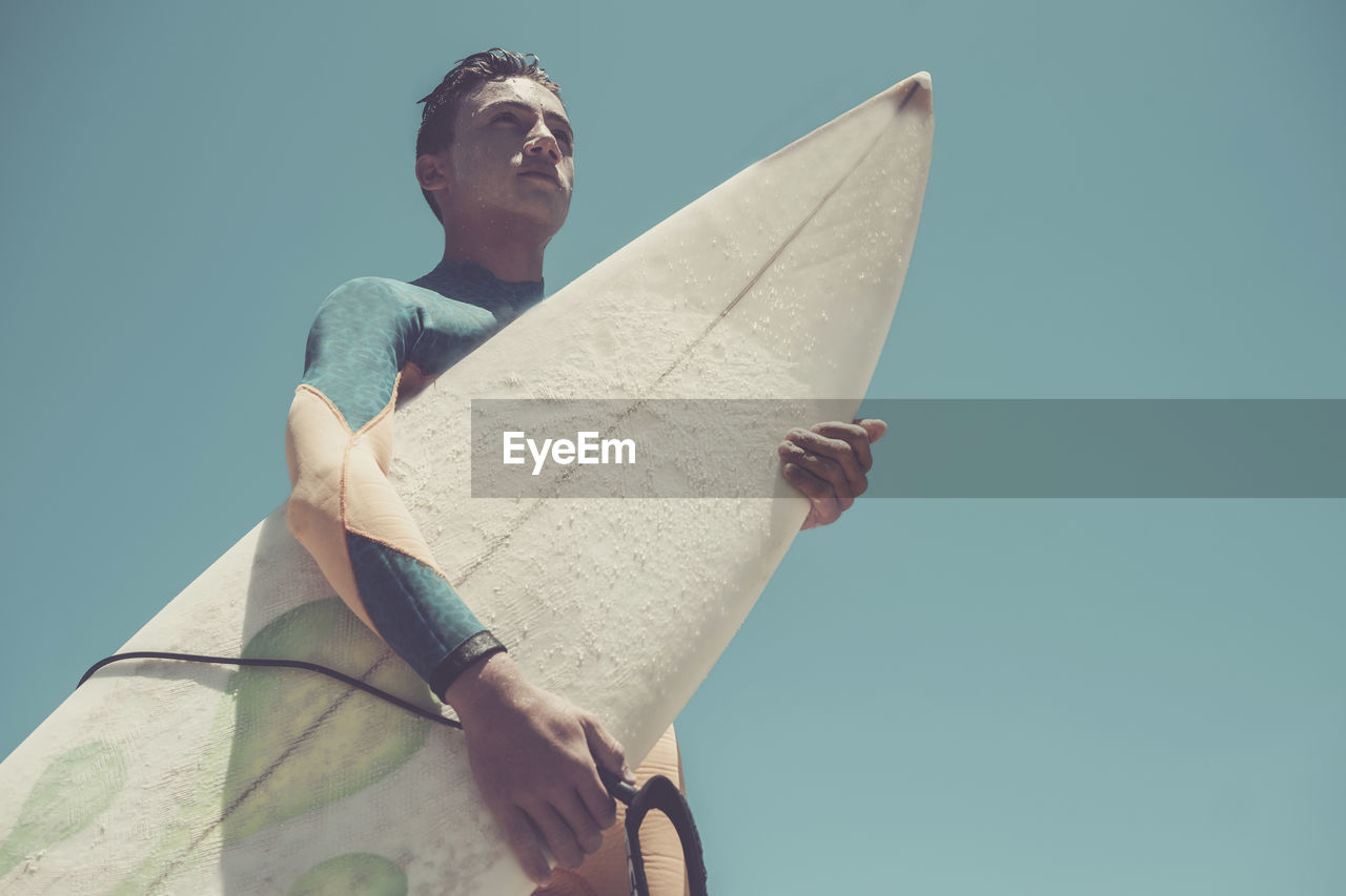 Low angle view of teenage boy holding surfboard against clear blue sky