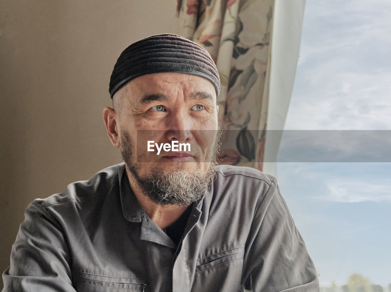 Senior asian man with beard wearing skullcap sitting by window and looking into distance. 