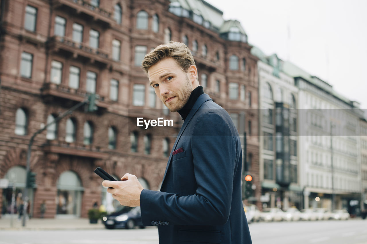 Portrait of businessman with smart phone standing against building in city