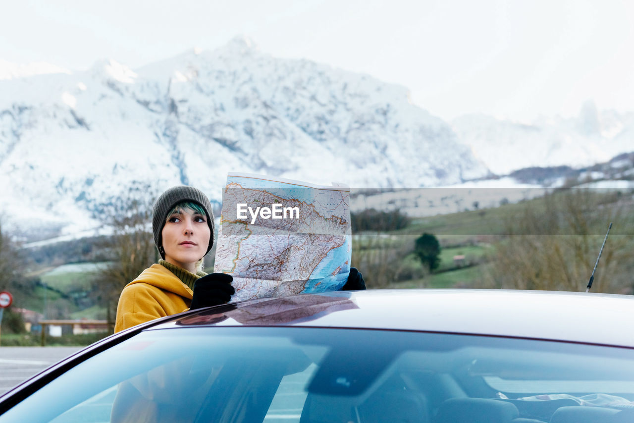 Attractive young female in outerwear standing with territory map near modern car during road trip in snowy mountainous terrain on cold winter day