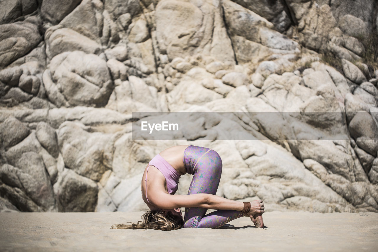 Woman practicing yoga at beach against rocks