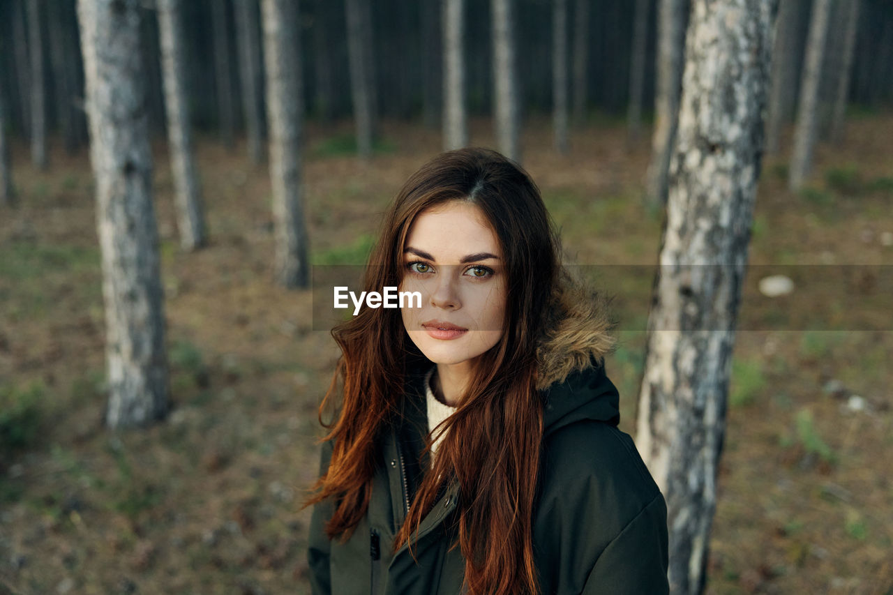 Portrait of young woman standing by tree trunk in forest