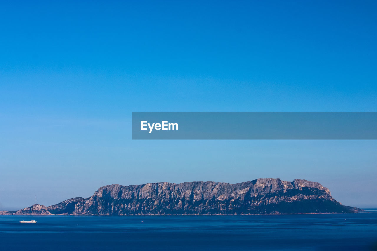 Scenic view of rock mountain by sea against clear blue sky