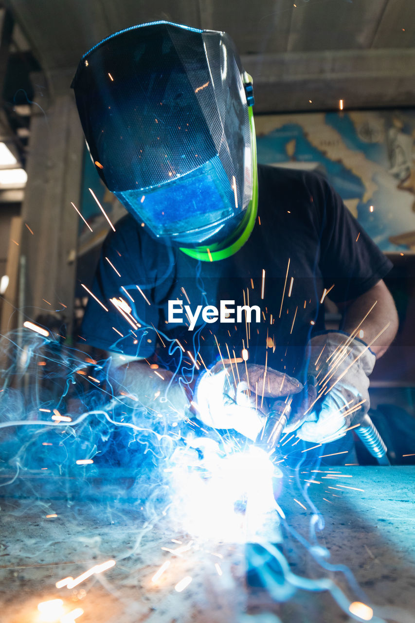 Unrecognizable male master in protective helmet welding metal piece at workbench in workshop