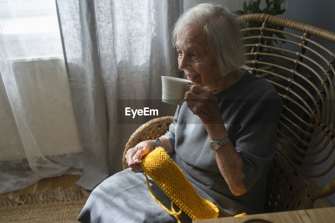 Senior woman having tea holding knitting wool on chair at home