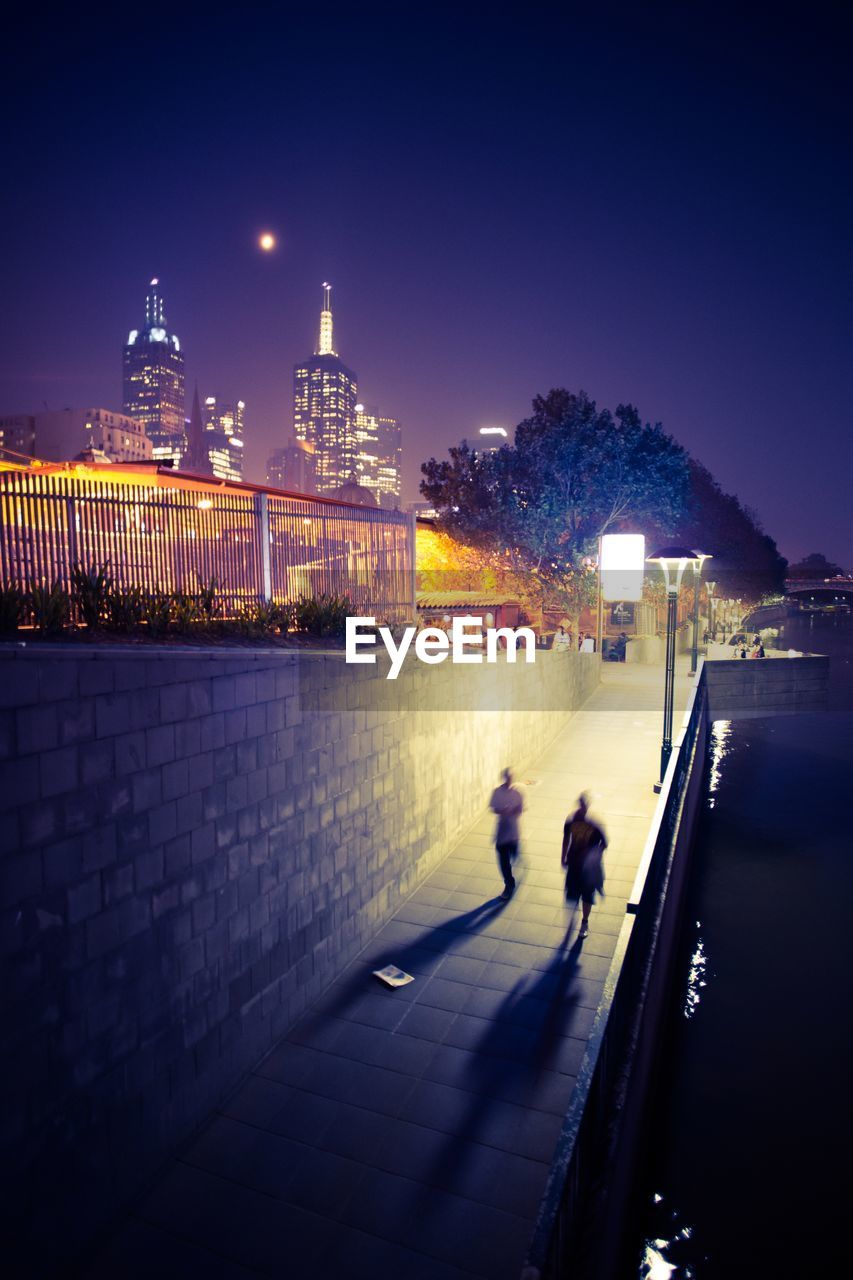 Blurred people walking on walkway by river against sky at night