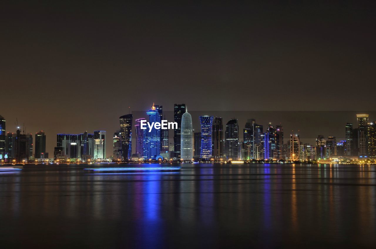 Illuminated modern buildings by bay against sky at night