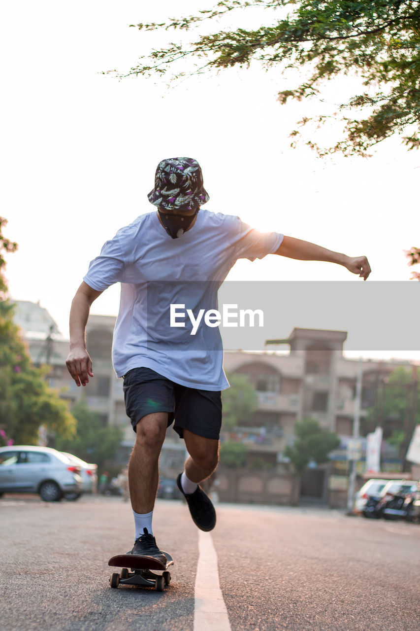A young adult skateboarding on an empty street while wearing a protective mask during sunrise.