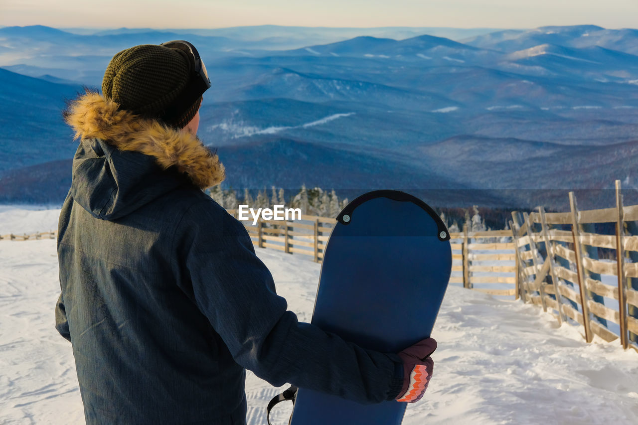 Rear view of man holding snowboard standing on snow covered land