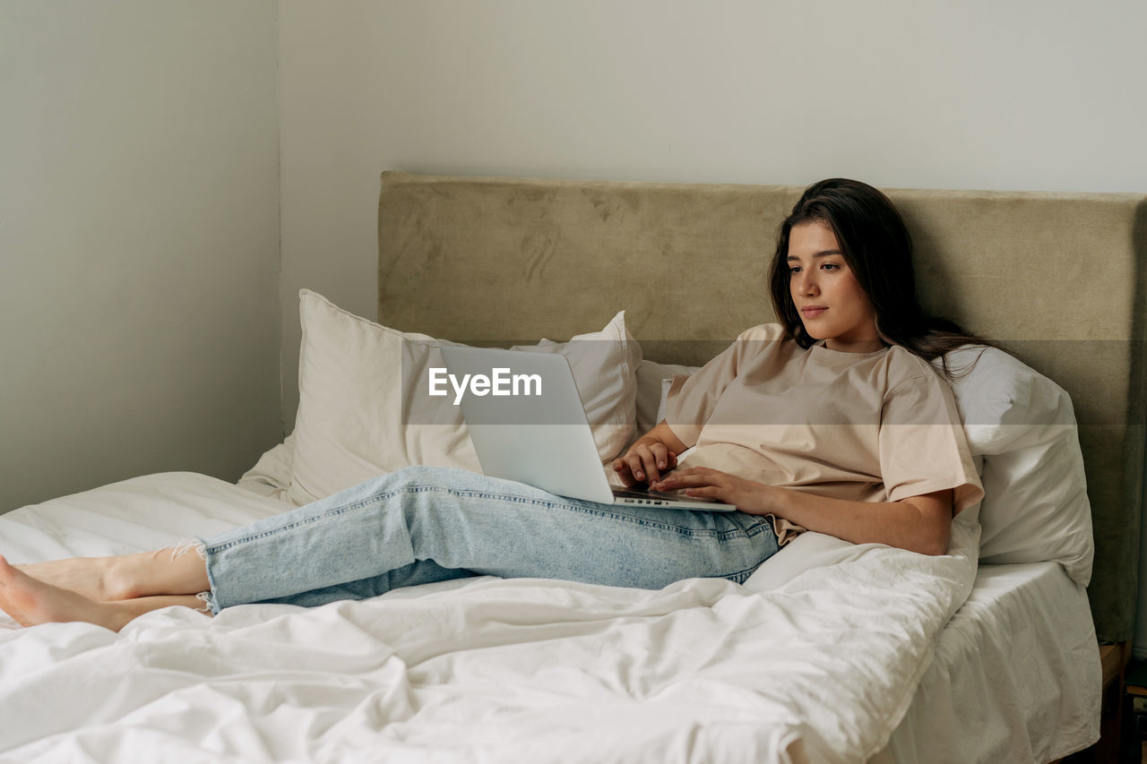 Young brunette freelancer in t-shirt and jeans lying on bed working on laptop