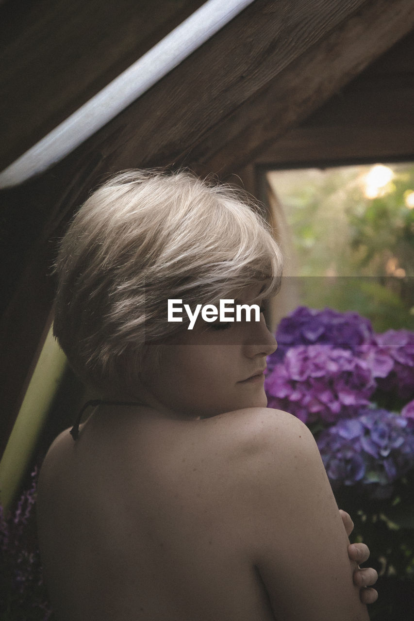 Rear view of shirtless young woman standing by flowers