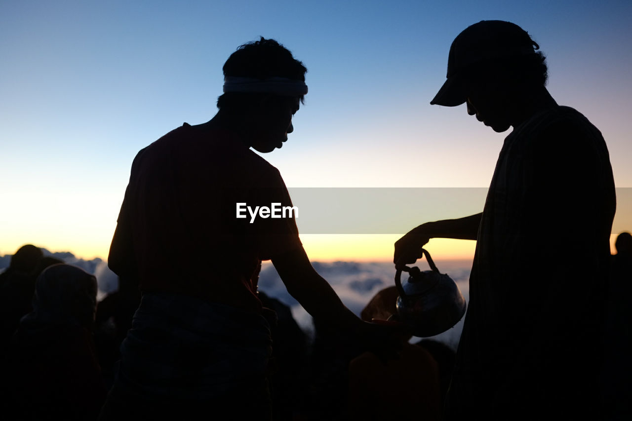 Silhouette man pouring coffee for hiker against sky during sunset