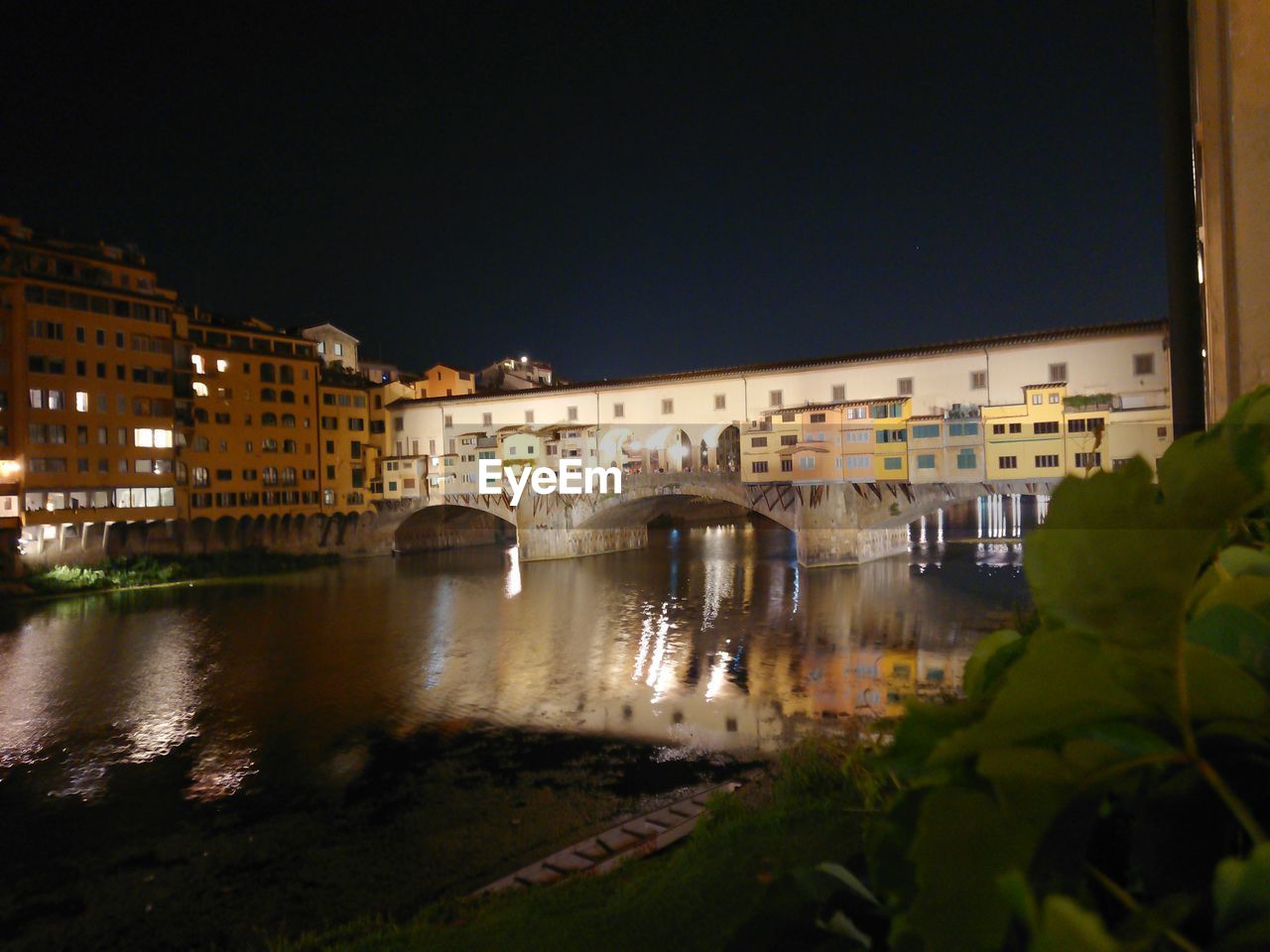 ARCH BRIDGE OVER RIVER AMIDST BUILDINGS AT NIGHT