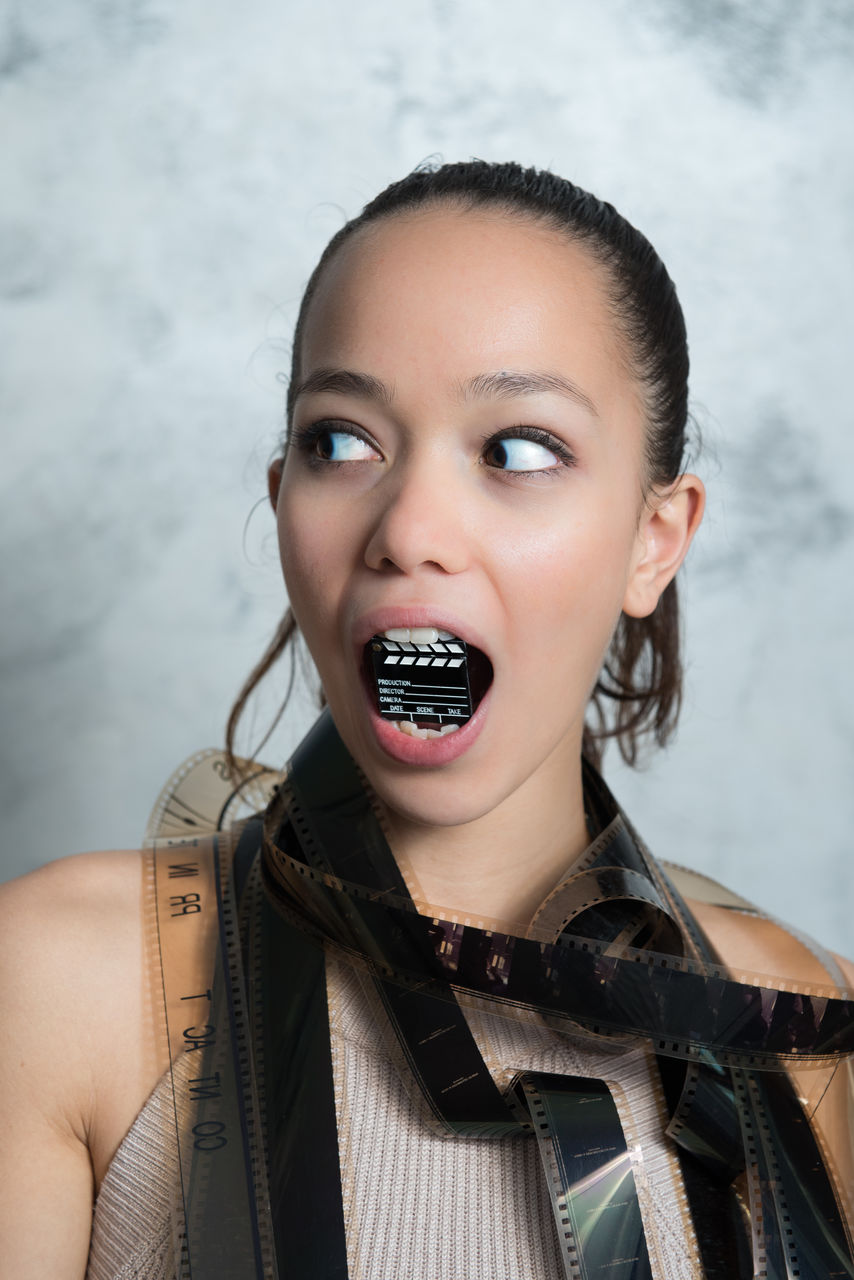 Young woman holding small clapperboard in mouth with reels around neck against wall