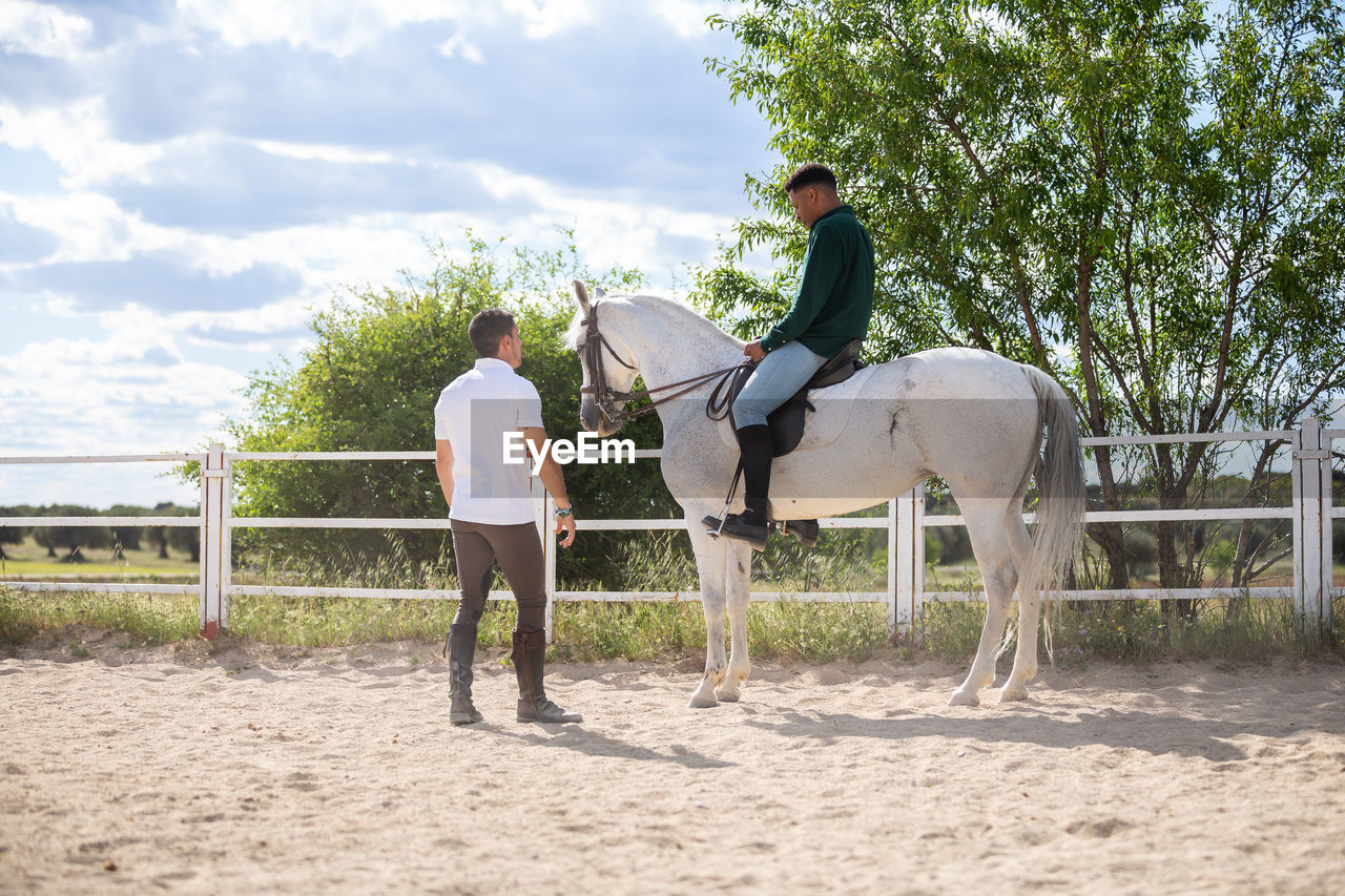 Man standing by friend sitting on horse in pen