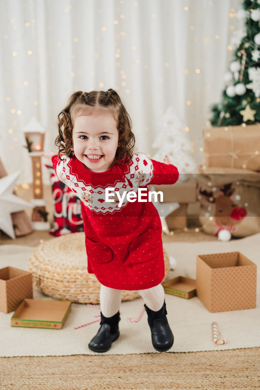 childhood, child, christmas, celebration, happiness, smiling, holiday, gift, emotion, full length, christmas tree, one person, portrait, cheerful, looking at camera, indoors, cute, front view, toddler, female, clothing, decoration, christmas decoration, tree, red, standing, box, innocence, fun, tradition, domestic room, person, joy, women, winter, baby, enjoyment, home interior, nature, domestic life, positive emotion, holding, event, excitement, lifestyles, living room, anticipation, plant