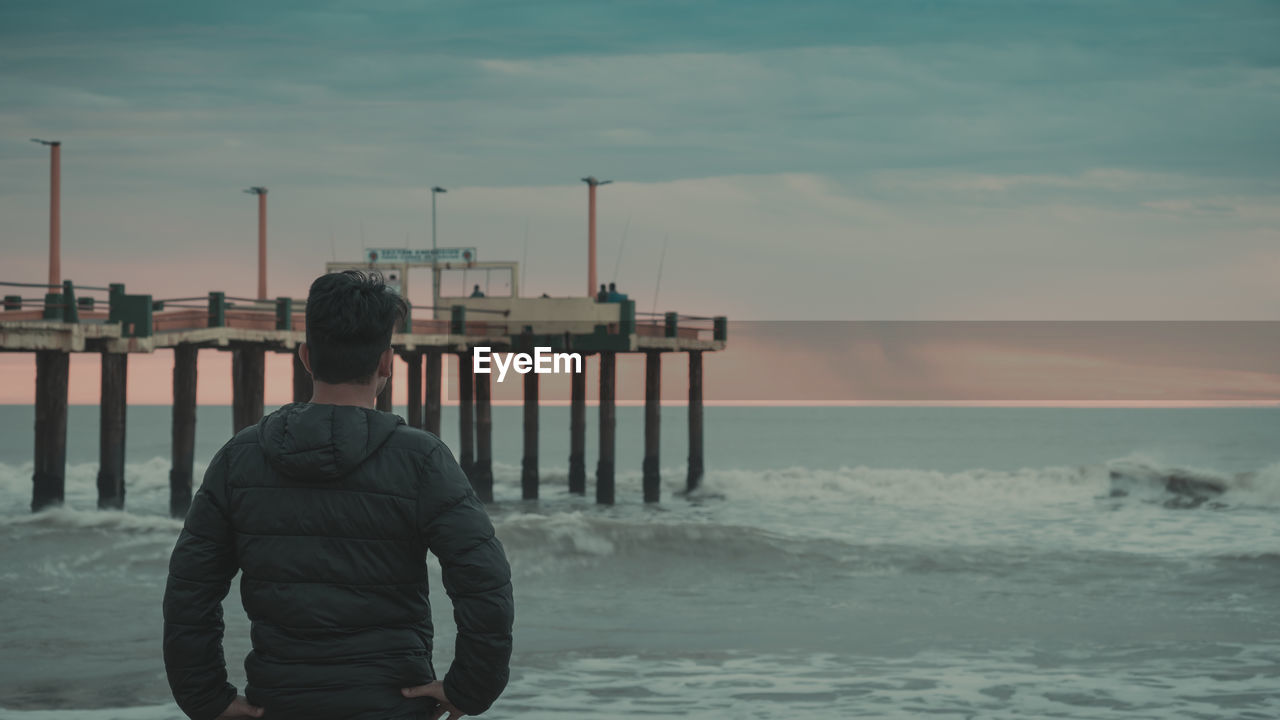 REAR VIEW OF MAN STANDING ON BEACH AGAINST SKY