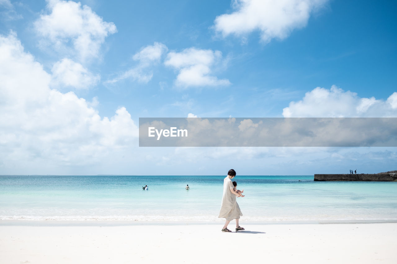 Woman carrying baby while walking at beach against sky