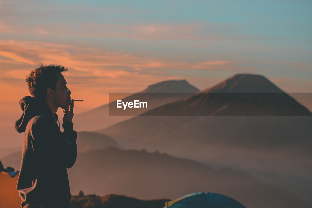 Side view of man smoking by mountains during sunrise
