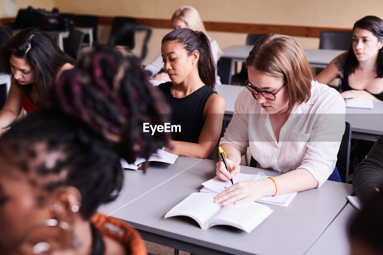 High angle view of female student writing in book while sitting with classmates in classroom