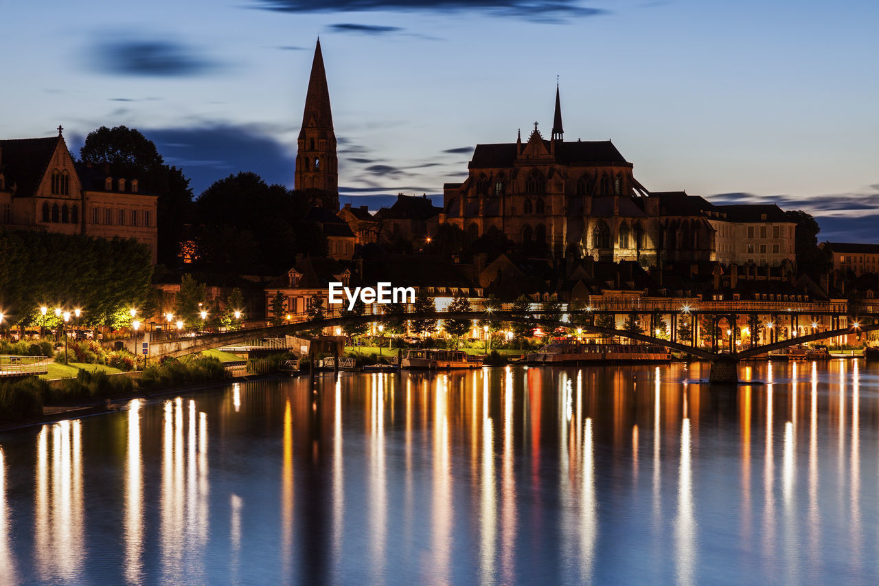 illuminated buildings by river against sky at night