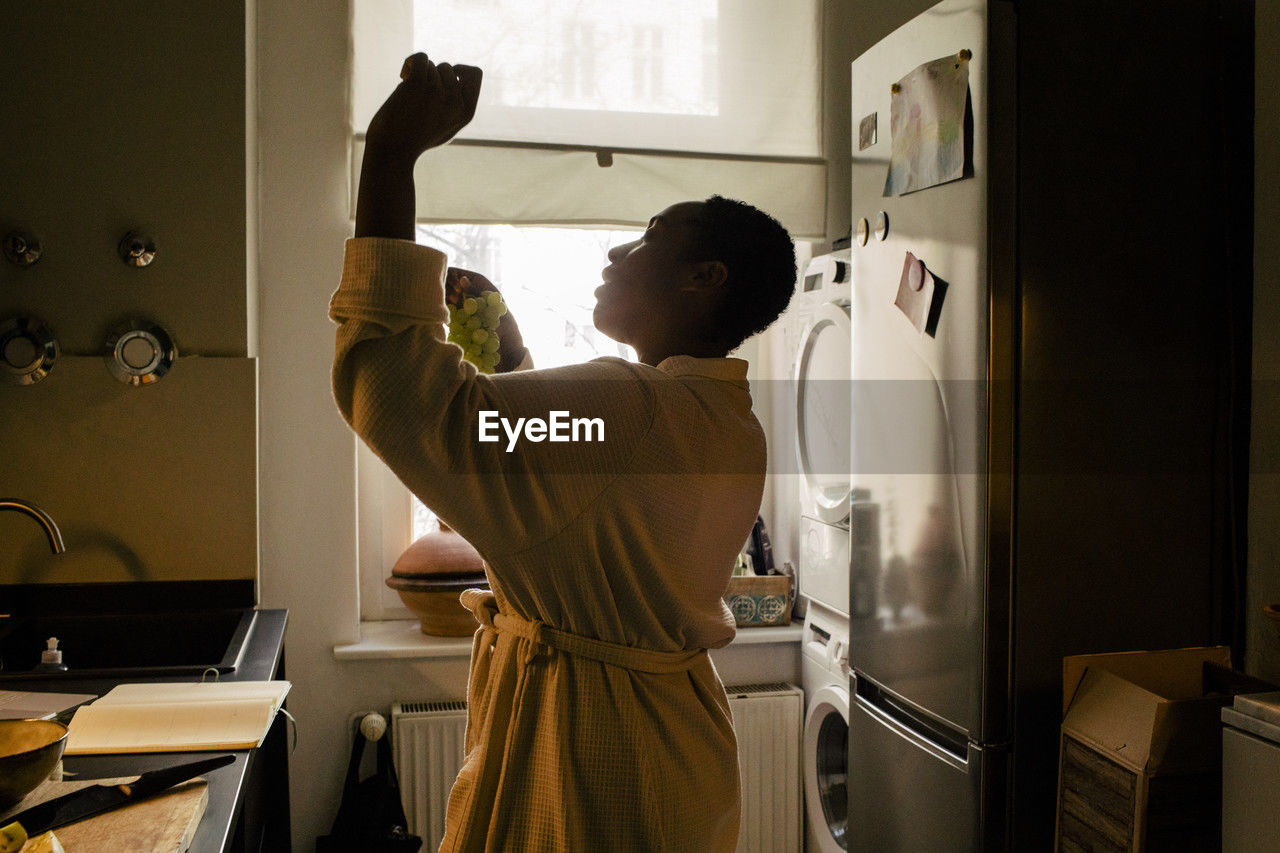 Carefree young woman holding grapes dancing in kitchen at home