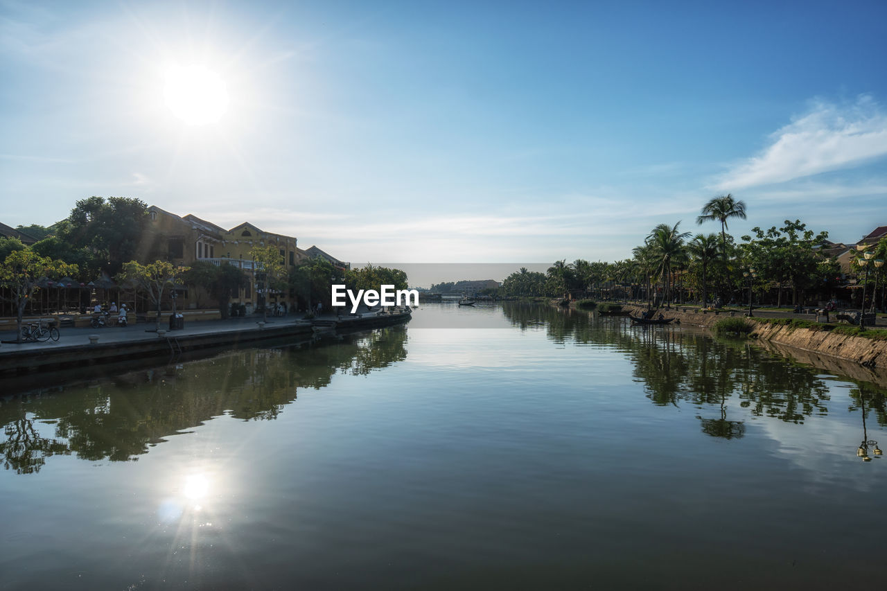 reflection, water, sky, river, nature, body of water, sun, tree, beauty in nature, tranquility, sunlight, architecture, scenics - nature, morning, plant, no people, tranquil scene, built structure, cloud, dusk, waterway, travel destinations, outdoors, sunbeam, blue, travel, building, environment, lens flare, building exterior, day, idyllic, tourism, landscape, waterfront, reservoir