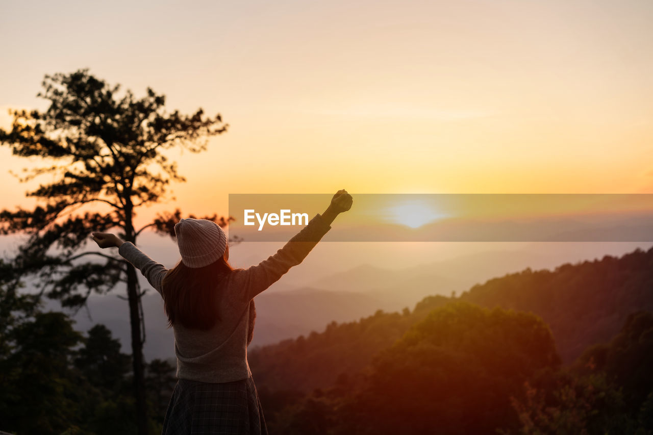 Rear view of woman with arms raised standing against sky during sunrise
