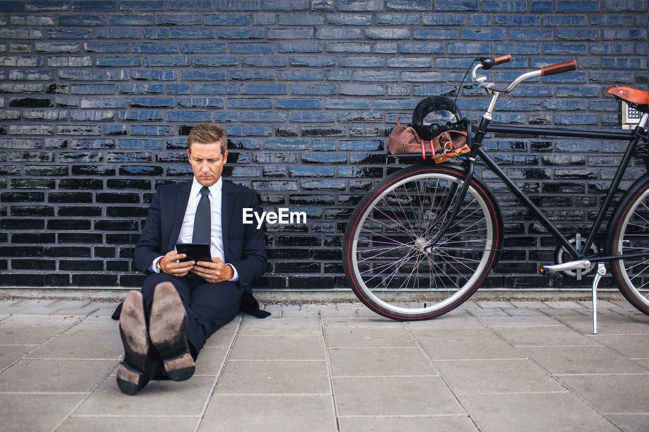 Full length of businessman using digital tablet while sitting on sidewalk by bicycle