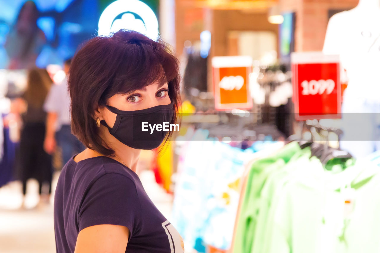 A woman in a black protective mask in a shopping mall among hangers with things during the pandemic.