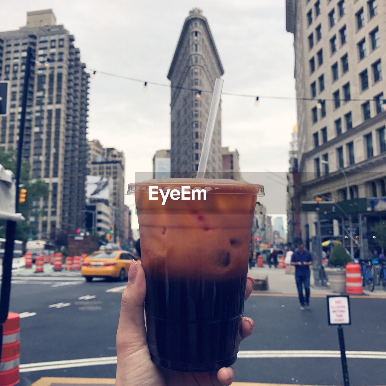 CLOSE-UP OF HAND HOLDING DRINK AGAINST BUILDINGS