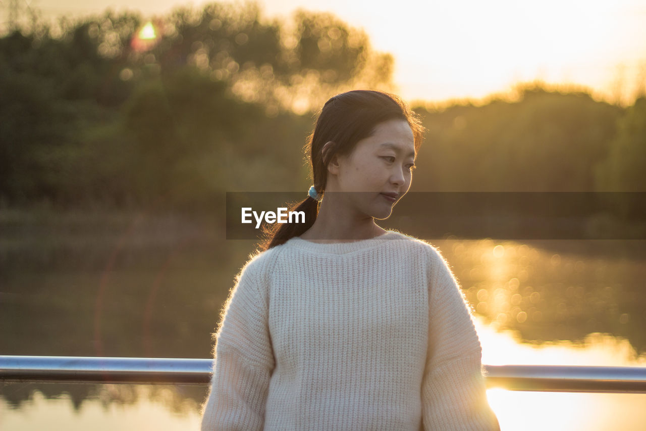 Thoughtful young woman standing against lake during sunset