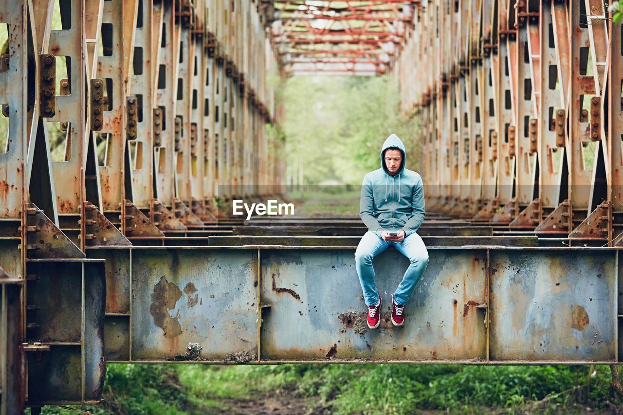 Full length of young man using phone while sitting on abandoned bridge