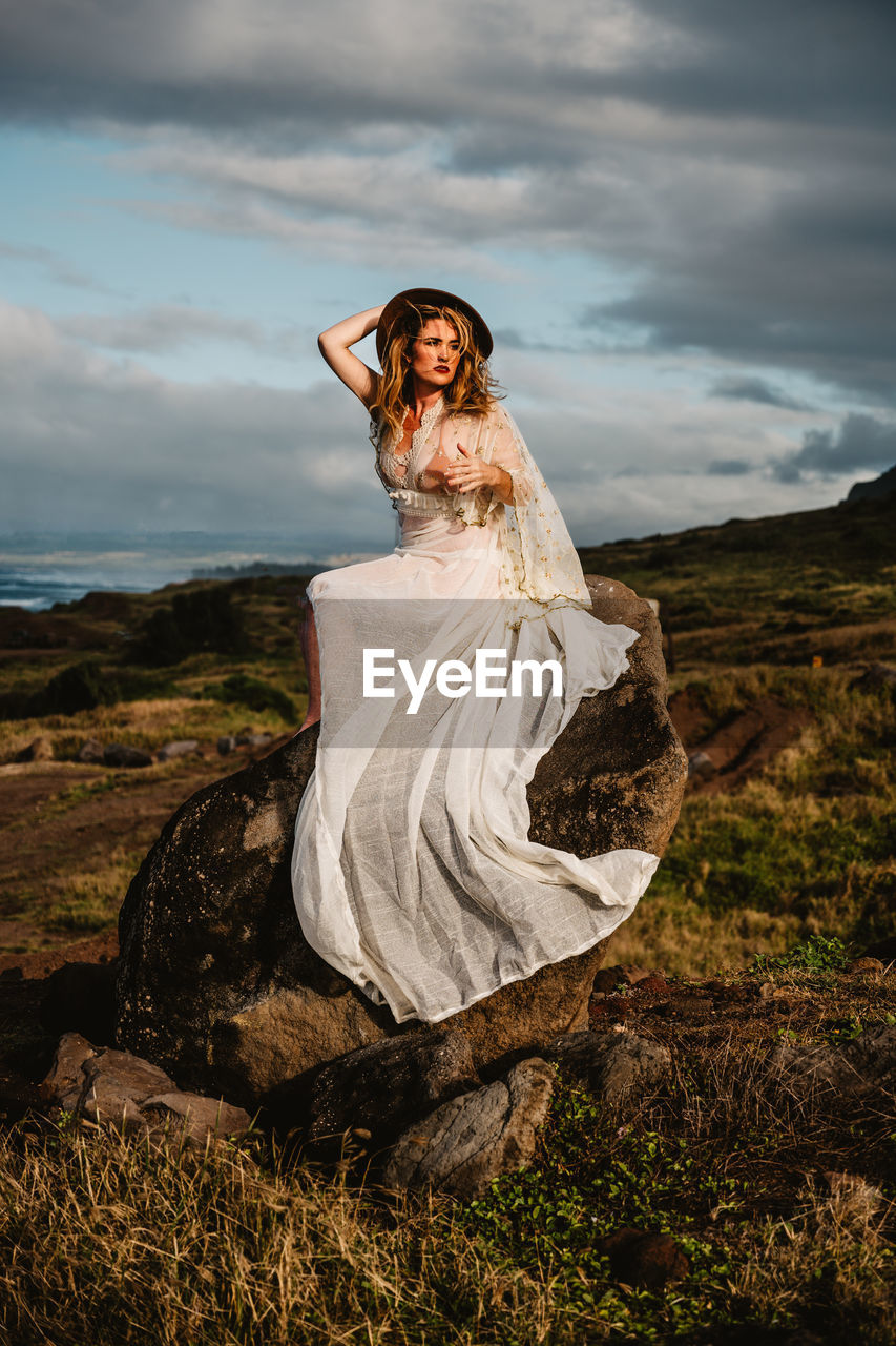 Woman wearing white dress while sitting on rock against cloudy sky