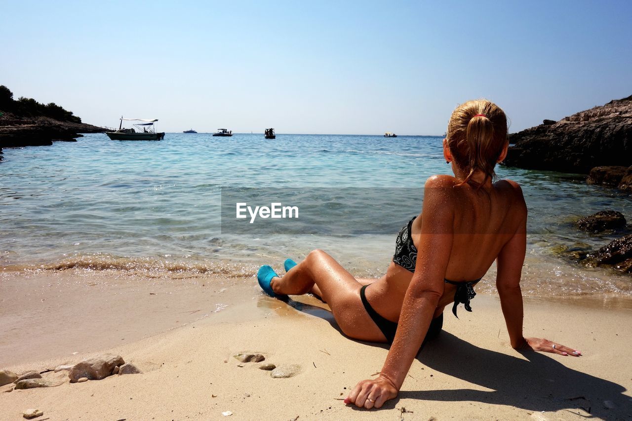 REAR VIEW OF SHIRTLESS MAN SITTING ON SHORE