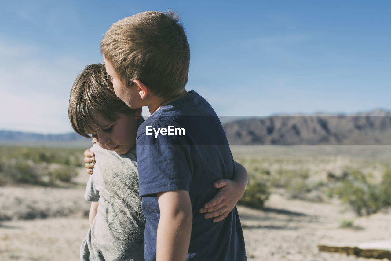 Boy kissing brother while standing at joshua tree national park against sky during sunny day