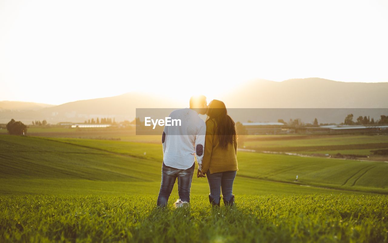 Rear view of couple kissing while standing on field against sky during sunset