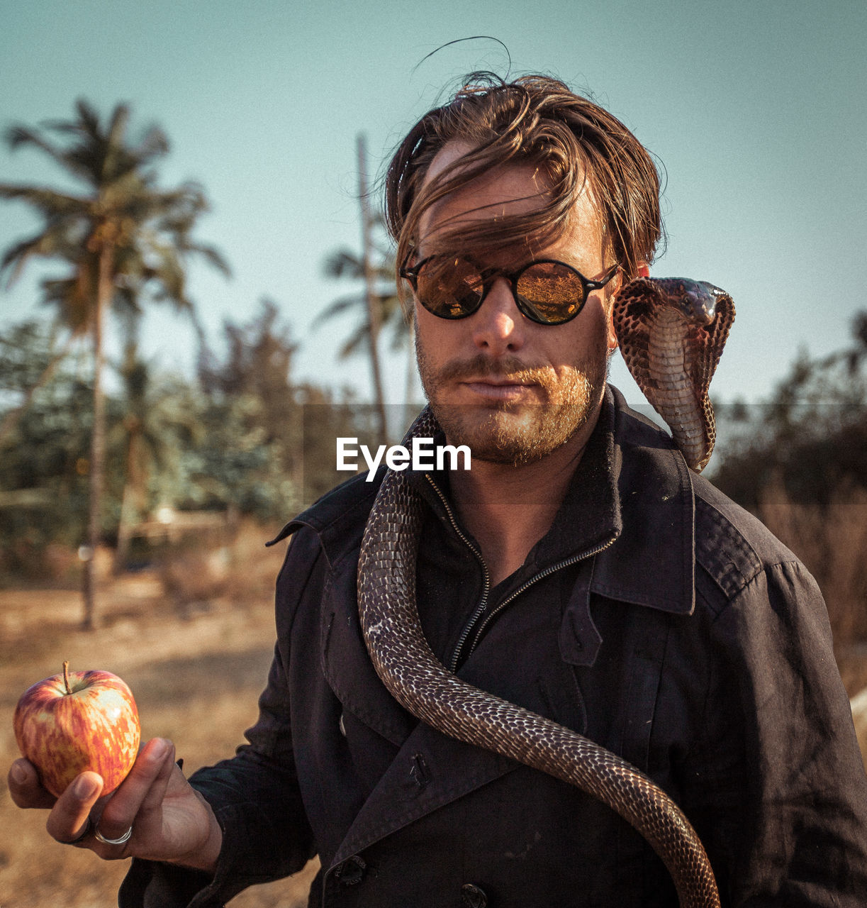 Portrait of mature man with snake holding apple standing against sky