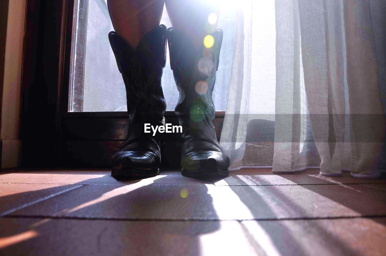 Low section of person standing in cowboy boots by window at home