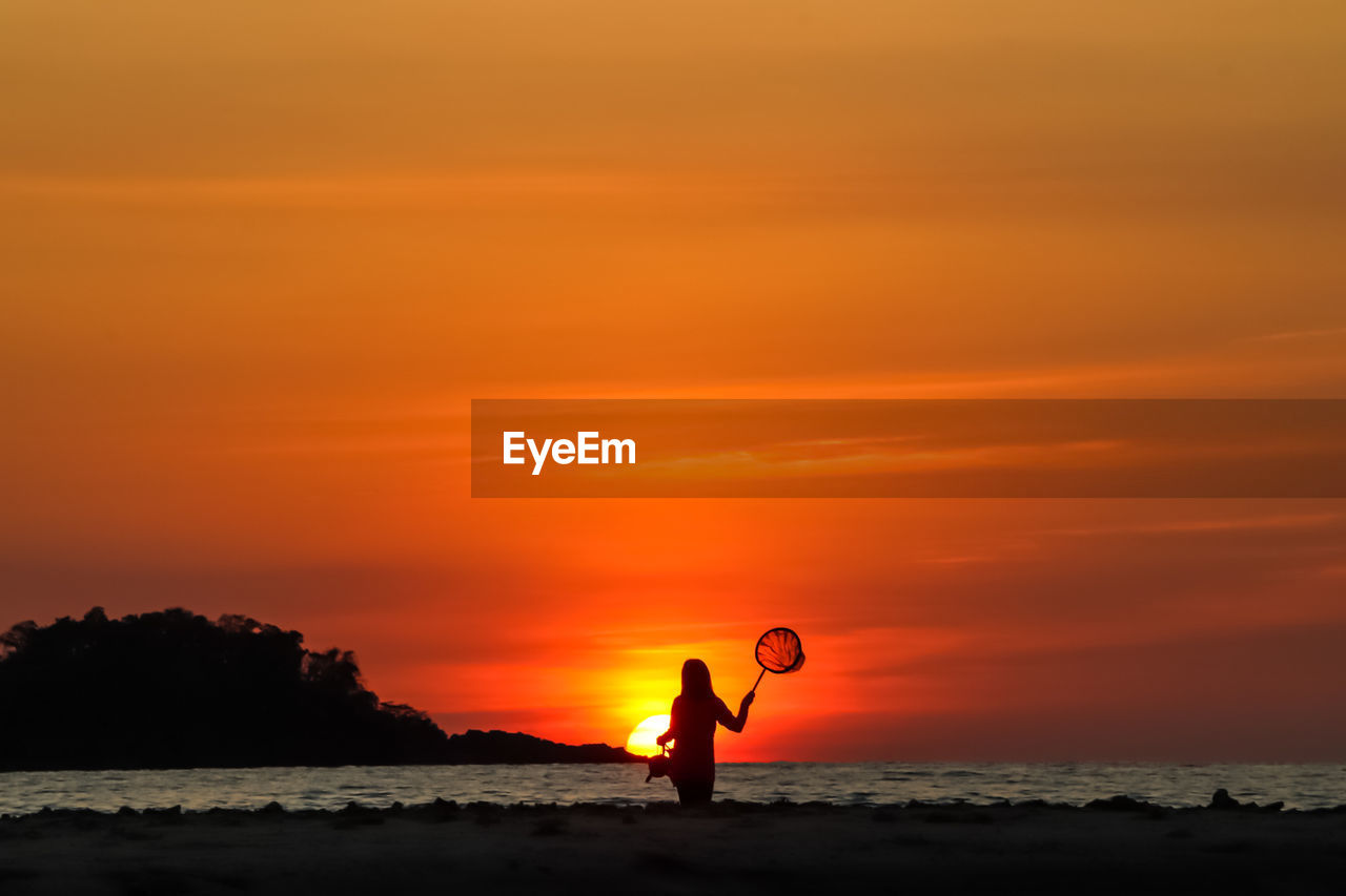 SILHOUETTE PERSON STANDING ON BEACH AGAINST ORANGE SKY